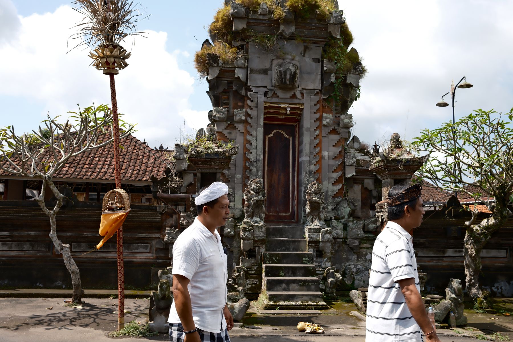 Two men walk past a grass-topped stone gate and penjor pole